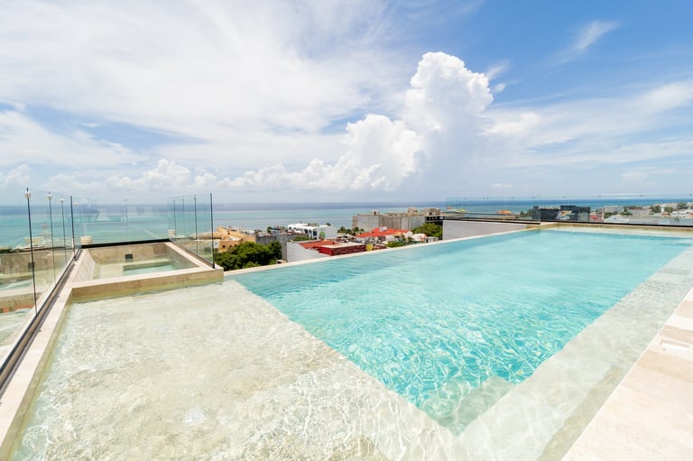 Rooftop infinity pool with turquoise water overlooking coastal town and ocean under blue cloudy sky