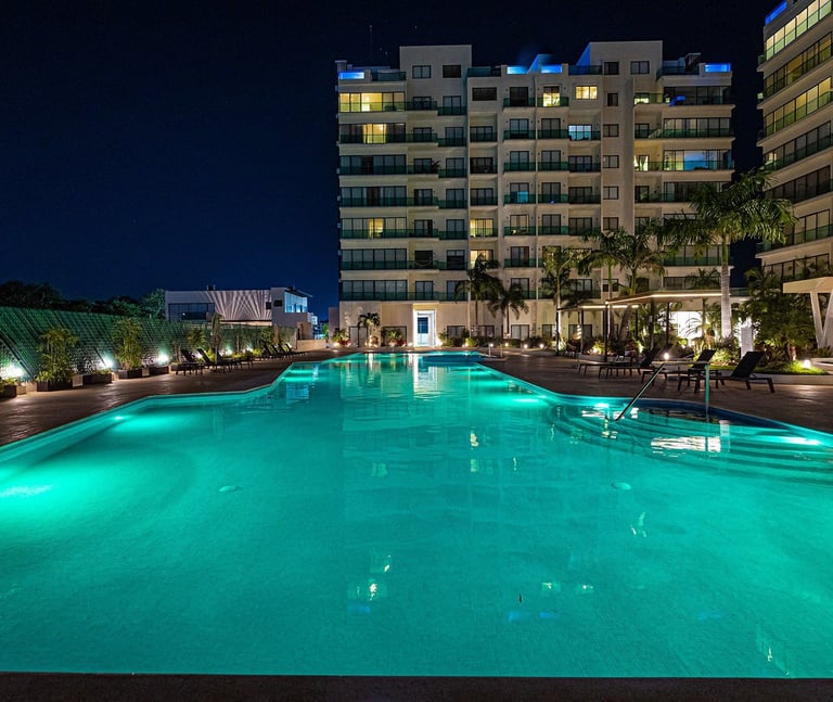 Illuminated turquoise swimming pool at night surrounded by modern residential buildings and tropical landscaping