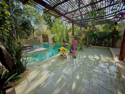 Tropical indoor pool area with turquoise water, pergola overhead, pink hanging flowers, and lush green plants surrounding the space