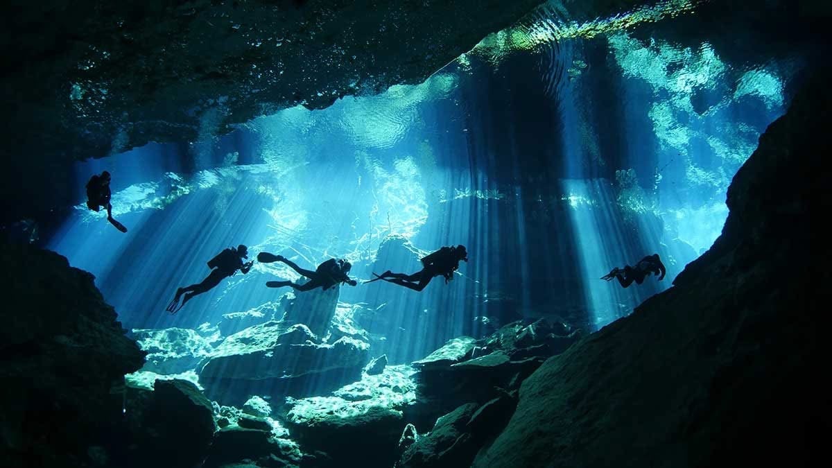 Multiple scuba divers exploring an underwater cave with sunlight filtering through the opening above