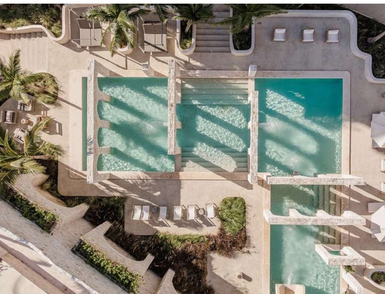 Aerial view of multiple turquoise swimming pools surrounded by white concrete deck, palm trees, and beige buildings
