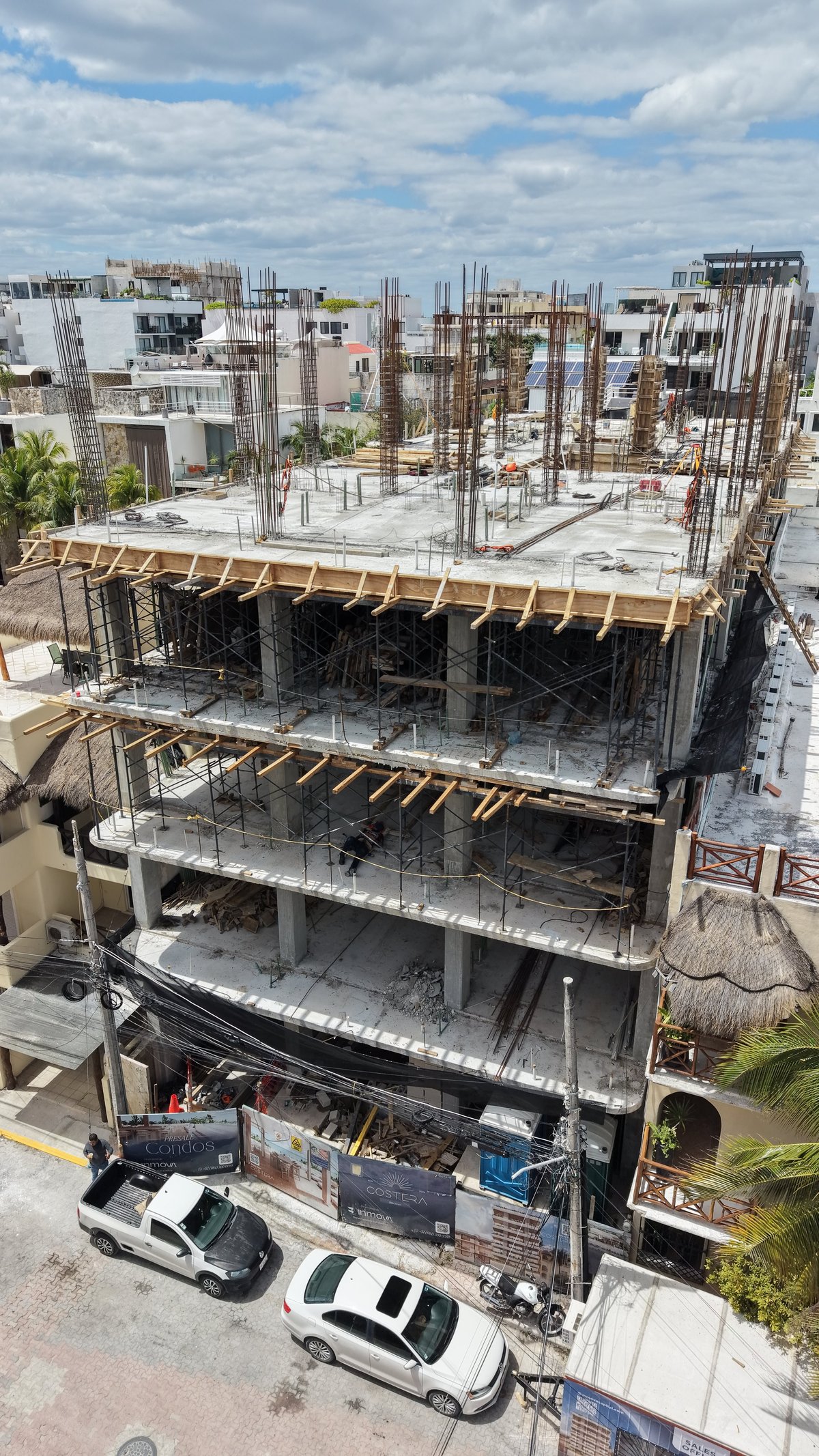 Multi-story building under construction with concrete floors, steel reinforcement bars, and wooden formwork on upper levels, surrounded by parked vehicles and other buildings