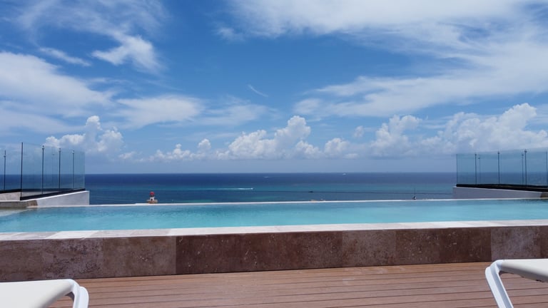 Modern infinity pool overlooking the ocean with blue sky and clouds, wooden deck and sailboats visible on horizon
