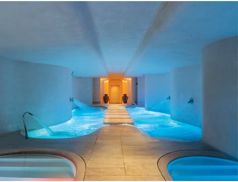 Modern spa interior with glowing blue water features on either side of a wooden floor corridor leading to an illuminated orange doorway