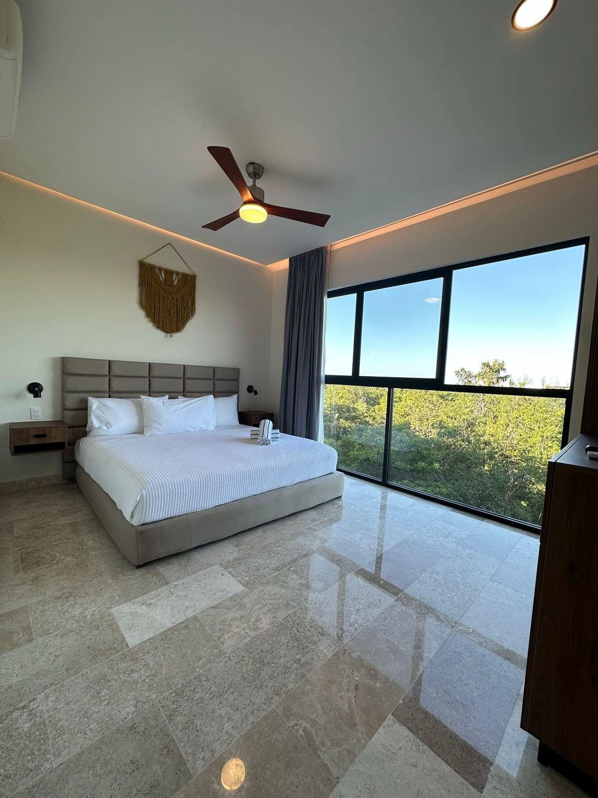 Modern bedroom with gray upholstered bed, polished tile flooring, ceiling fan, and large windows overlooking greenery