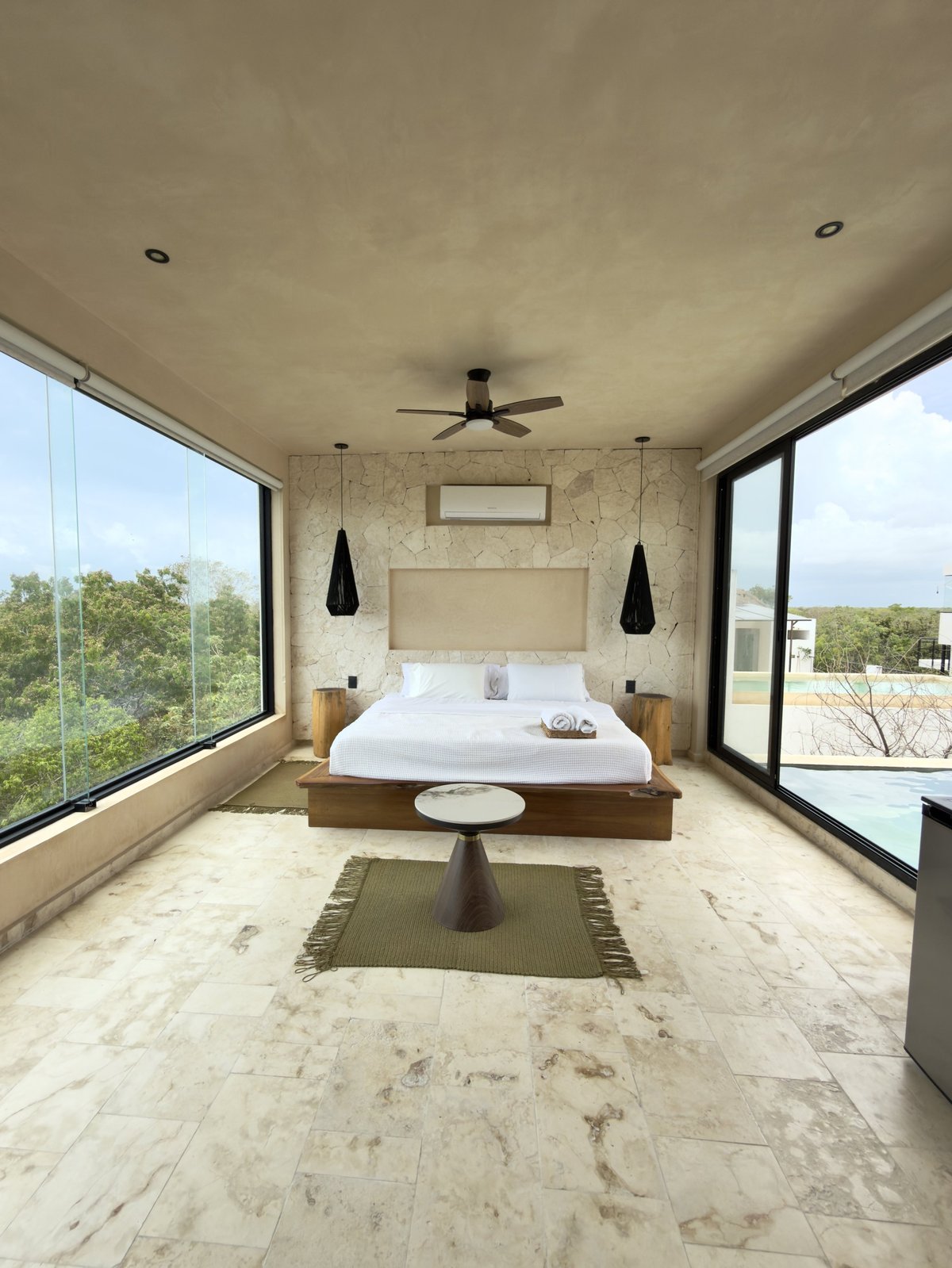 Modern bedroom with large windows, marble flooring, wooden bed with white linens, pendant lights, and ceiling fan overlooking landscape
