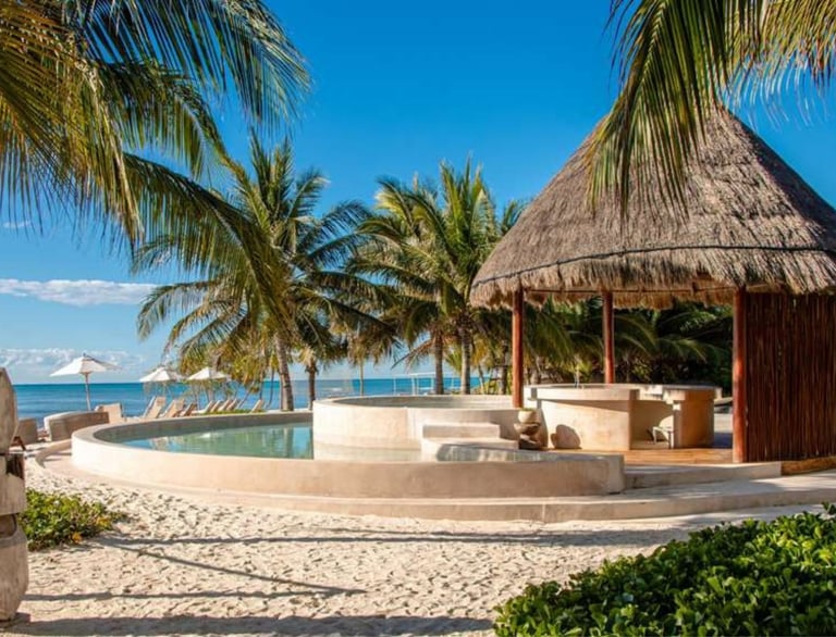 Luxury tropical beach resort with thatched-roof cabana, swimming pool, palm trees, and ocean view under clear blue sky