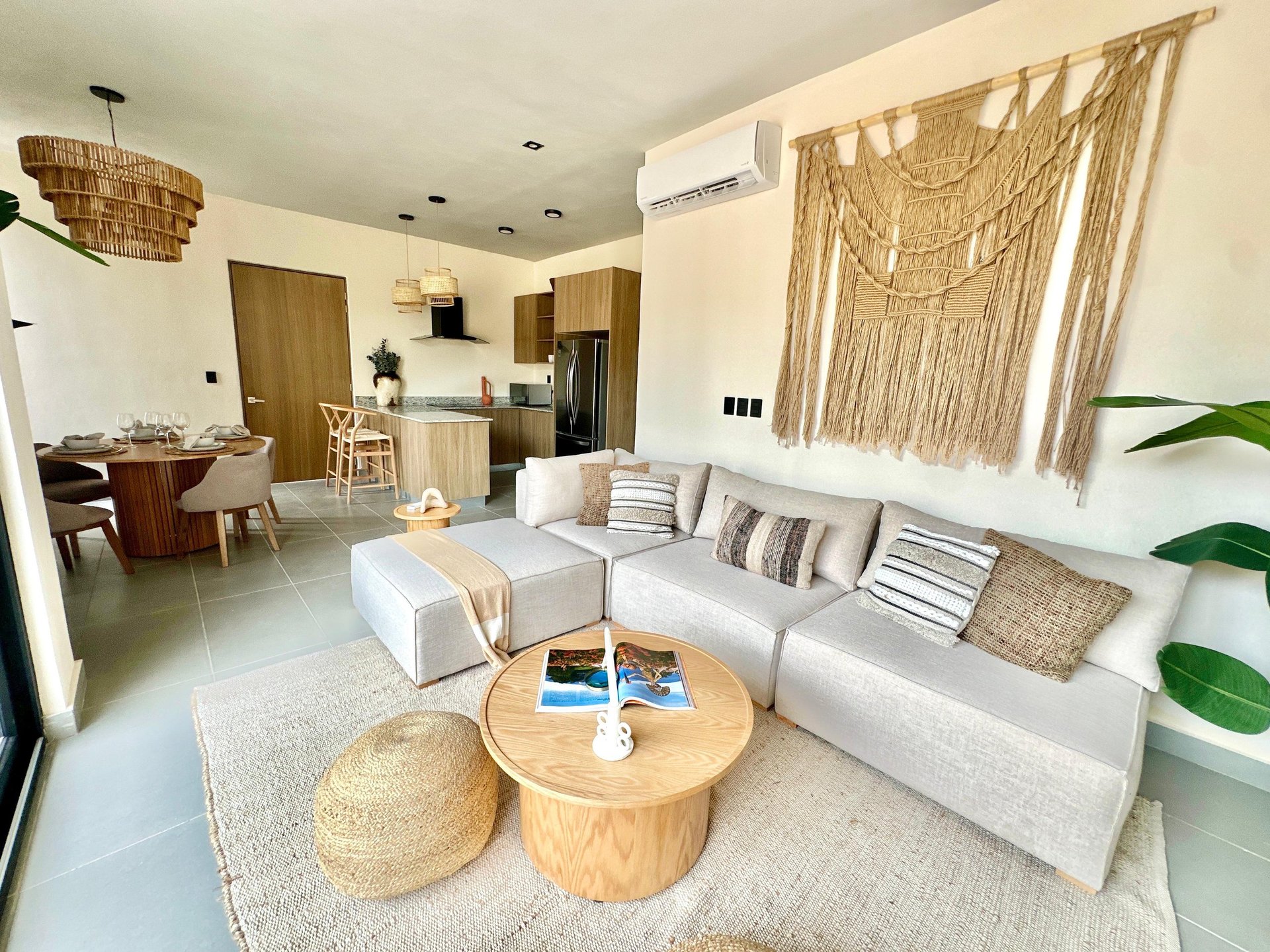 Modern open-concept apartment interior with white sofa, wooden coffee tables, macramé wall hanging, and connected kitchen and dining areas