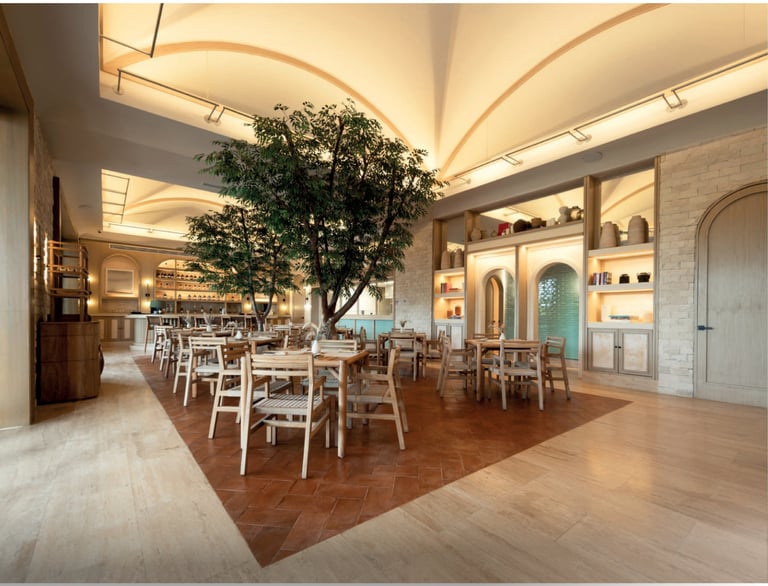 Modern restaurant interior with large green tree centerpiece, white chairs, warm ceiling lighting, wooden herringbone floor, and built-in shelving along walls