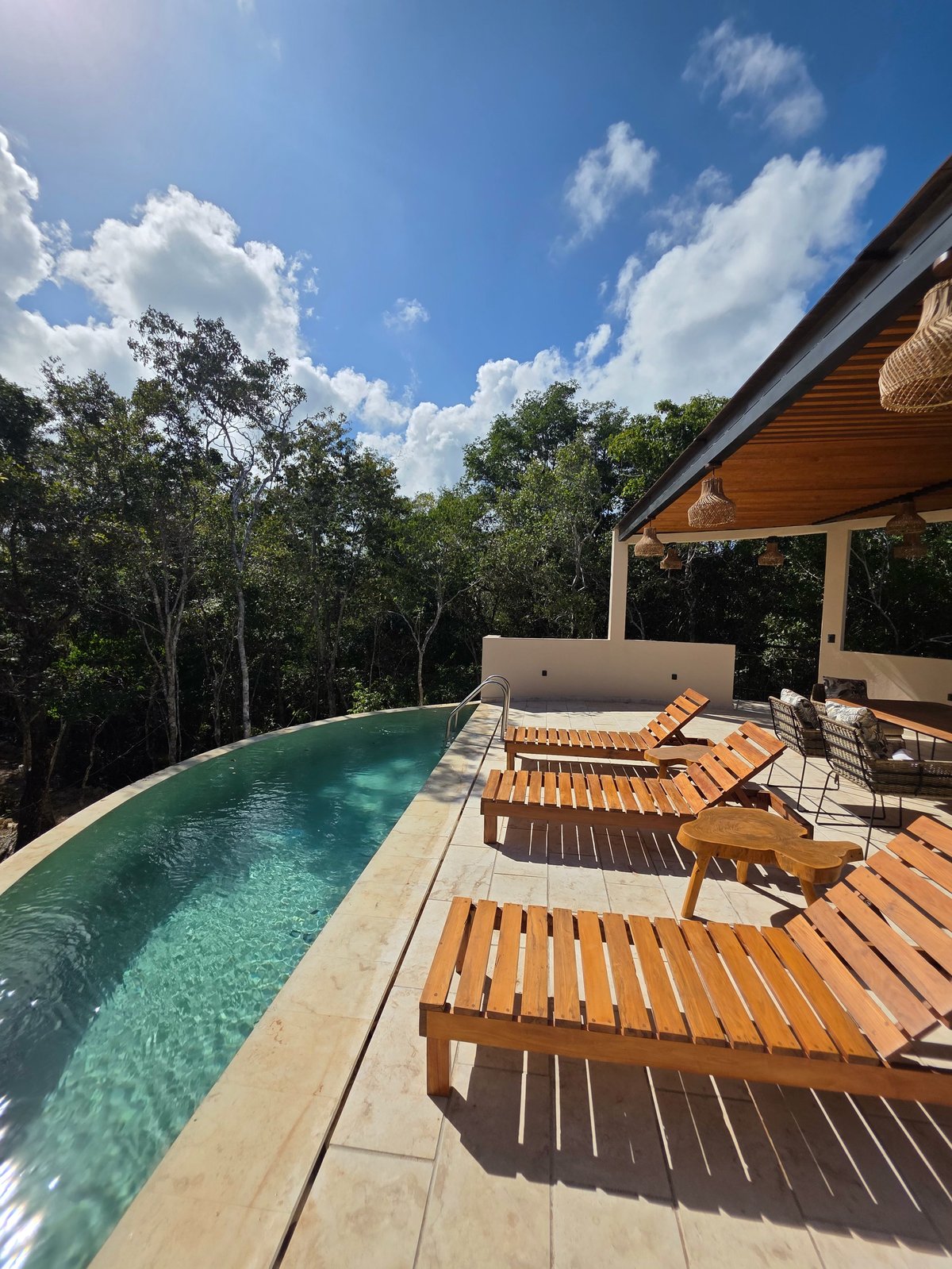 Luxury resort infinity pool with wooden lounge chairs on a deck, wooden pergola, forest backdrop, and blue sky with clouds