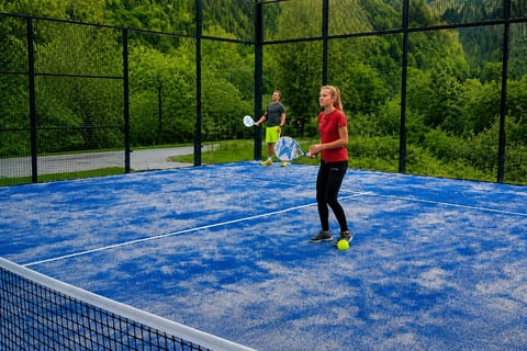 Child in red shirt playing tennis on blue outdoor court with green trees in background and protective netting surrounding the court
