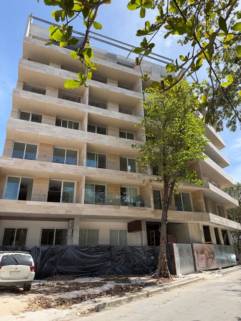 Beige concrete apartment building under construction with black tarps covering the lower level, large tree beside it, and clear blue sky