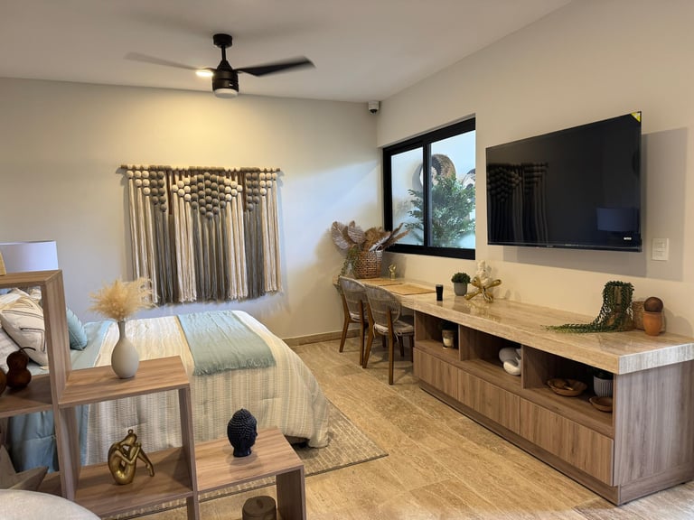 Modern bedroom with wooden furniture, TV mounted on beige wall, macramé wall hanging, and natural light from window