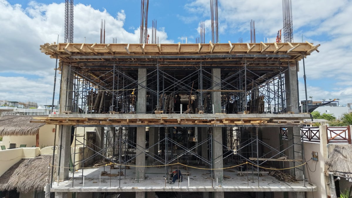 Multi-story building under construction with concrete columns, steel reinforcement, wooden formwork, and scaffolding against cloudy sky