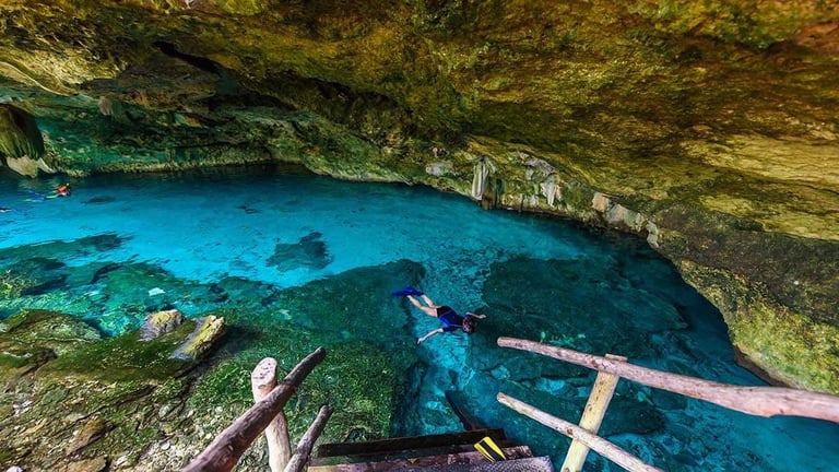 Snorkeler in turquoise cenote pool beneath rocky cave ceiling with fallen logs in foreground