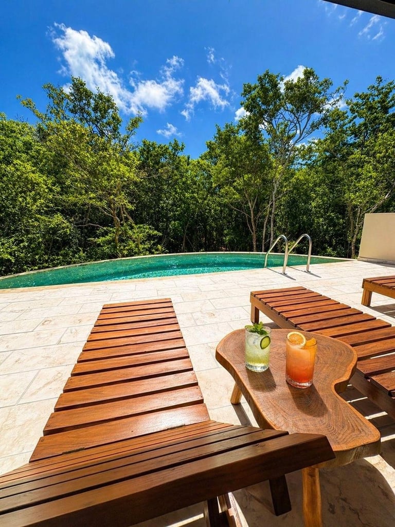 Luxury backyard pool area with wooden lounge chairs, turquoise pool, white deck, tropical trees, and drinks on a wooden tray
