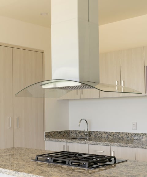 Modern kitchen with curved glass range hood, granite countertop, gas stovetop, and light wood cabinetry