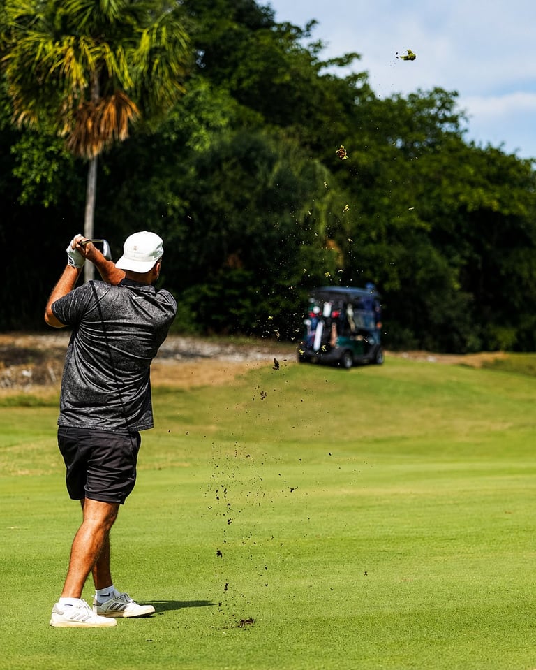 Golfer in black shirt and white cap mid-swing on lush green course with palm trees and golf cart in background