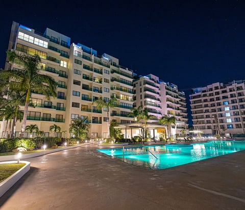 Illuminated apartment complex at night with turquoise swimming pool, palm trees, and multi-story residential buildings