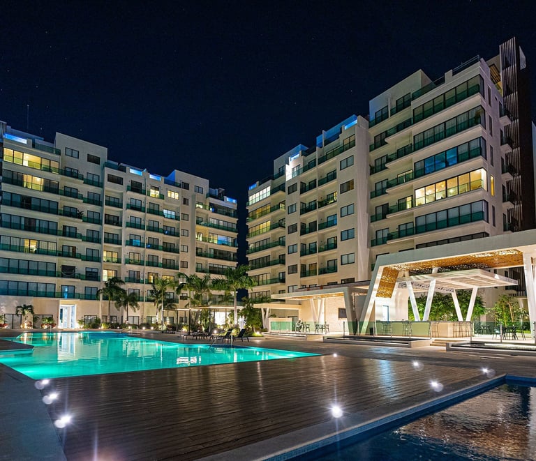 Nighttime view of a resort with illuminated swimming pool, modern apartment buildings, and tropical plants