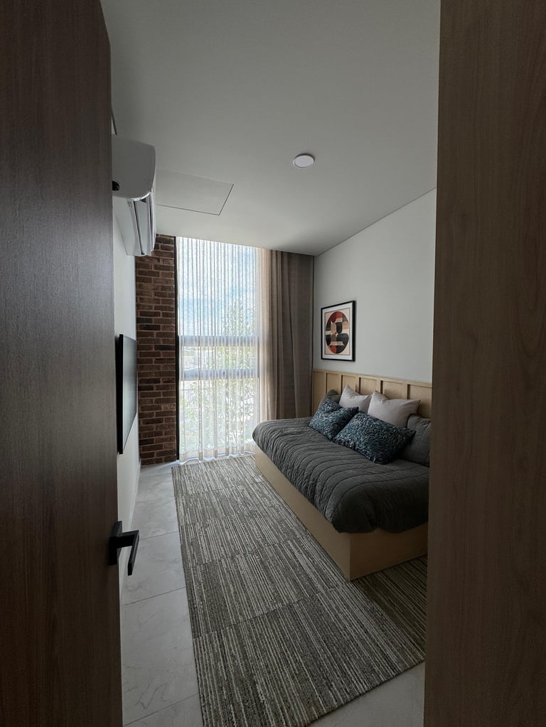 Modern bedroom viewed from doorway with queen bed, brick accent wall, floor-to-ceiling window with sheer curtains