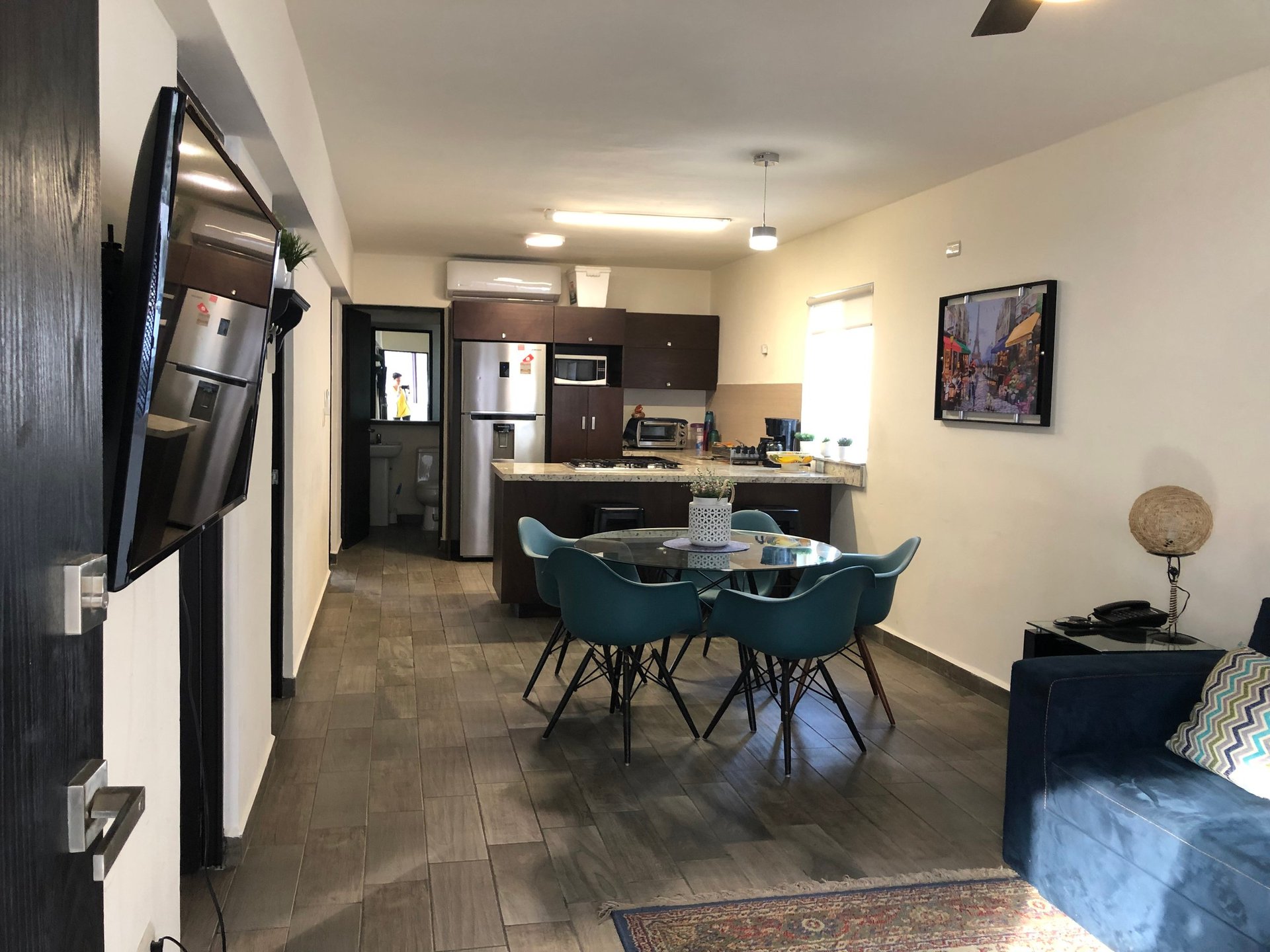 Modern open-concept apartment with blue dining table, kitchen, and living room with mounted TV and blue seating