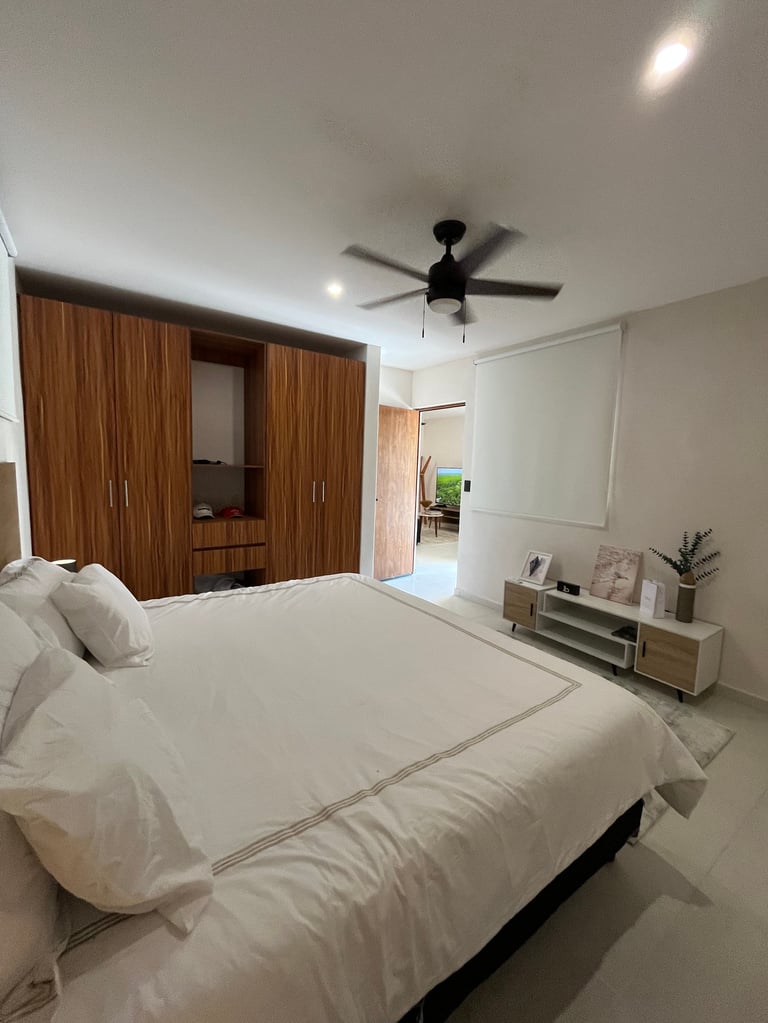 Modern bedroom with white bedding, wooden wardrobe, ceiling fan, recessed lighting, and minimalist decor