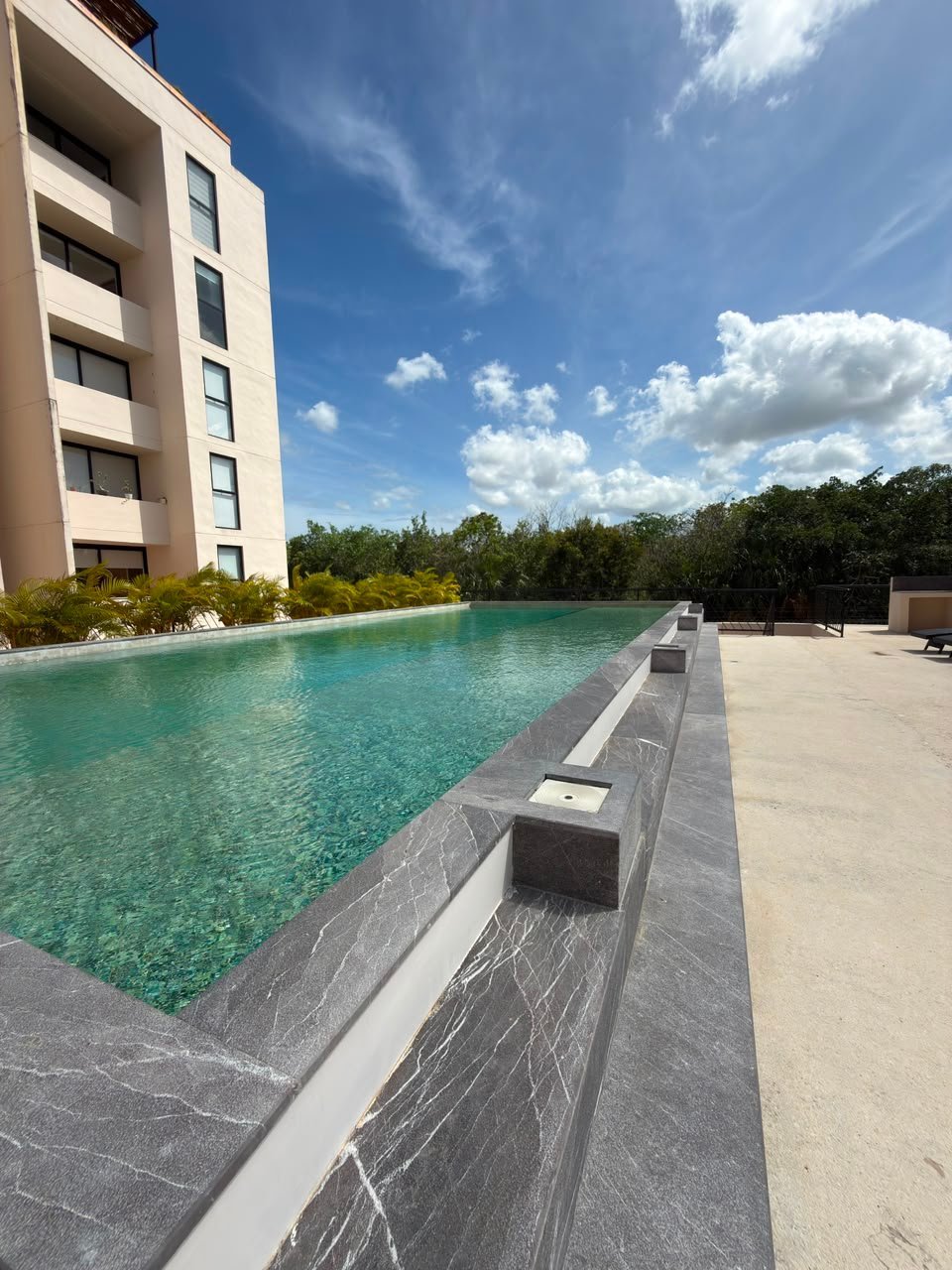 Modern apartment building with turquoise pool, concrete deck, and tropical vegetation under blue sky