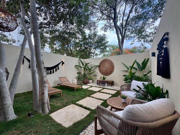 Serene outdoor courtyard with lounge chairs, potted plants, stepping stones, hammock, and white walls surrounded by trees
